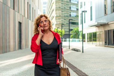 Kıdemli yönetici kadın hareketli, modern bir şehirde güvenle yürüyor, bir klasör ve şık bir çanta taşırken bir telefon görüşmesi yapıyor, profesyonelliği ve başarıyı somutlaştırıyor.