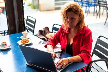 Üst düzey iş kadını laptopunu kullanıyor ve kahve ve kruvasanla kahvaltının tadını çıkarıyor. Uzaktan çalışıyor ve bağlantıda kalıyor.