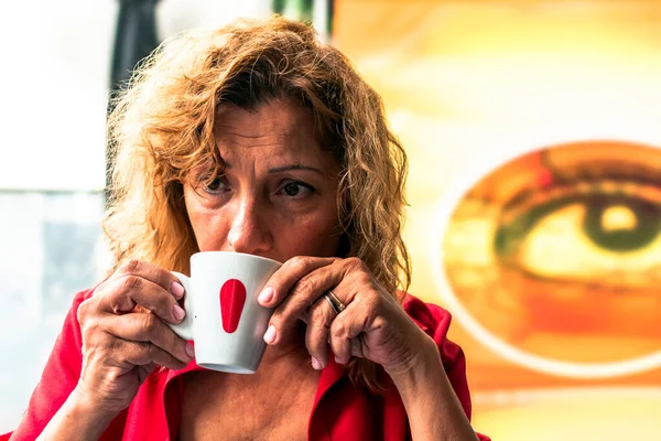 Kıdemli iş kadını, iş molasında bir fincan kahvenin keyfini çıkarıyor. Arkasındaki duvarı süsleyen eşsiz bir göz boyasıyla bir kafede oturuyor. İlginç ve sanatsal bir zemin oluşturuyor.