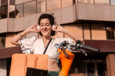 Mutlu genç bir kadın açık havada güneşli bir günün tadını çıkarıyor, elinde renkli alışveriş torbalarıyla kulaklıkla müzik dinliyor ve canlı bir şehirde bisiklet paylaşım hizmeti veriyor.