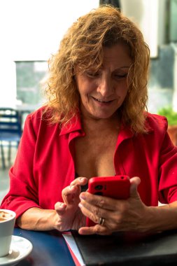 Kafe masasında akıllı telefon kullanan canlı kırmızı kıyafetli üst düzey yönetici bir kadın yanında bir fincan kahve, profesyonel bağlılığı ve modern iletişimi somutlaştırıyor.