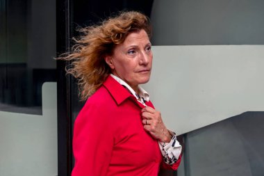 Başarılı üst düzey yönetici kadın kırmızı ceketini düzeltirken rüzgar saçlarını okşuyor, şehir ortamında şık, modern cam bir binanın yanında güvenle duruyor.