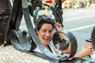 Güneş gözlükleriyle gülümseyen neşeli bir turist ve yeniden kullanılabilir bir kahve fincanı karmaşık dairesel desenler ve mozaik fayanslarla süslenmiş dekoratif metal bir kapının arkasında duruyor.