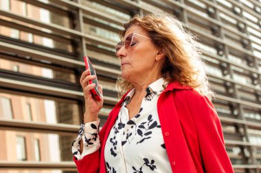 Akıllı telefon kullanan kıdemli iş kadını, modern bir ofis binasının yakınına sesli mesaj gönderiyor, iletişim, teknoloji ve iş dünyası gibi kavramları temsil ediyor.