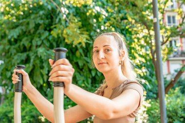 Yoğunlaşmış genç bir kadın, güneşli bir günde yeşil bir parkta spor aletlerini kullanarak kol kaslarını çalıştırıyor, sağlıklı yaşam tarzını ve formülünü teşvik ediyor.