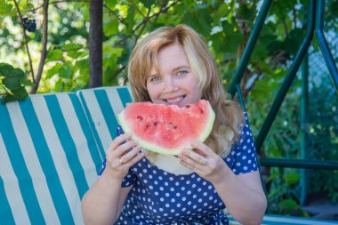 Genç bir kız yazlık alanında bir salıncak üzerinde oturan bir karpuz yiyor