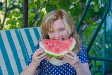 Genç bir kız yazlık alanında bir salıncak üzerinde oturan bir karpuz yiyor