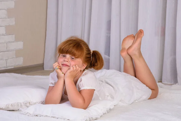 Little red-haired girl in a white dress lying on the floor, propping his chin with his hands — Stock Photo, Image