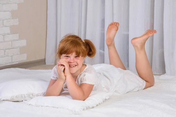 Little red-haired girl in a white dress is lying on the floor, propping her chin with her hands and laughing — Stock Photo, Image