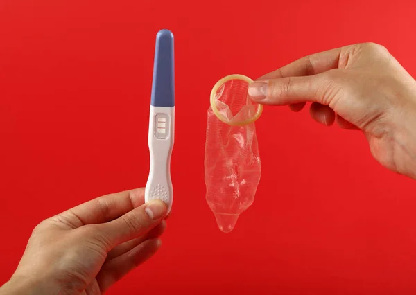 Close up woman hands holding positive (pregnant) pregnancy test and condom over red background with copy space, low angle view, personal perspective