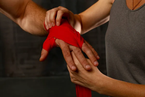 Close up woman trainer helps man boxer wrapping red hand wraps over wrists preparing for fight, over black background with copy space