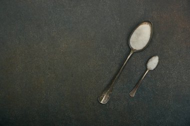 Düz açıda iki kaşık beyaz şeker ve deniz tuzu bulunur. Koyu grunge taş masa yüzeyinde fotokopi alanı ile birlikte. Üst görünümü tam üstte.