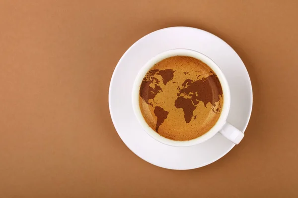 Beyaz bardağı çay tabağına kapat, dünya haritalı espresso kahvesi köpüklü krema ile doldur, arka planda fotokopi alanı olan kahverengi kağıt, üst görünümü tam üstte,