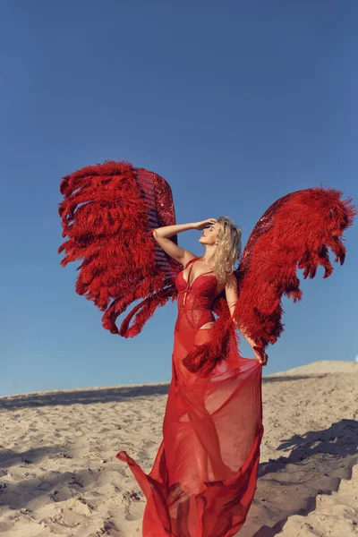 Mladá blondýnka s červenými křídly na písku — Stock fotografie