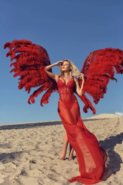 Mladá blondýnka s červenými křídly na písku — Stock fotografie