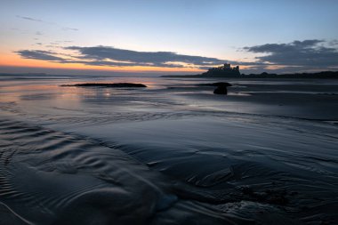 İngiltere 'nin kuzeydoğu kıyısındaki Bamburgh Şatosu, Northumberland' daki Bamburgh köyü. Dengesiz ve dramatik kıyı görüntüsü, gel-git dalgaları ve nehirler. Güneş doğduktan hemen sonra çekildi..