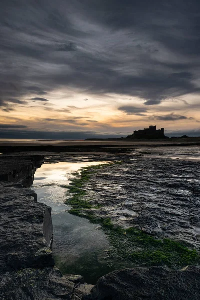 İngiltere 'nin kuzeydoğu kıyısındaki Bamburgh Şatosu, Northumberland' daki Bamburgh köyü. Karamsar ve karamsar bir kıyı görüntüsü ve heybetli karanlık gökyüzü. Güneş doğduktan hemen sonra çekildi.