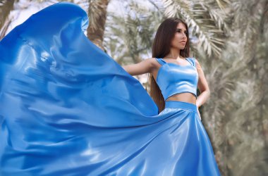 Harika esmer palmiye ağaçları ayakta uzun mavi ayrılmış elbise, sallama etek, kadın uzun saçlı, mükemmel bir vücut ve güzel yüzü, kalıcı makyaj, Güneş ışınları, yaz