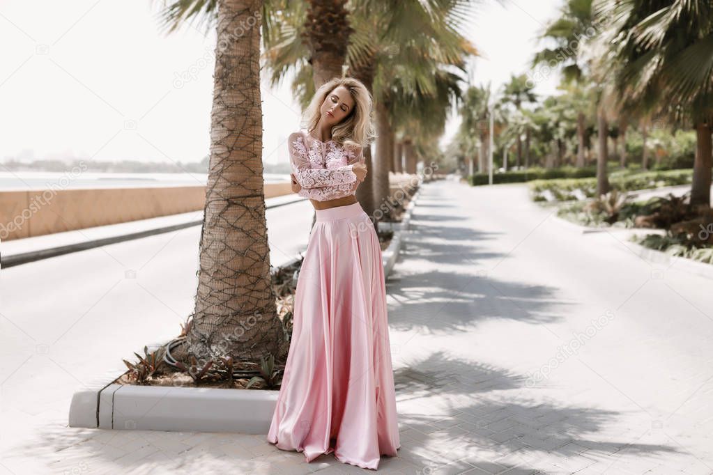 Shapely blonde next to the sea in Dubai, palm trees, hot, gorgeous dresses, summer sunny lifestyle fashion shooting, waving in the wind dress, calm and chilling out near the pool, hairstyle, makeup