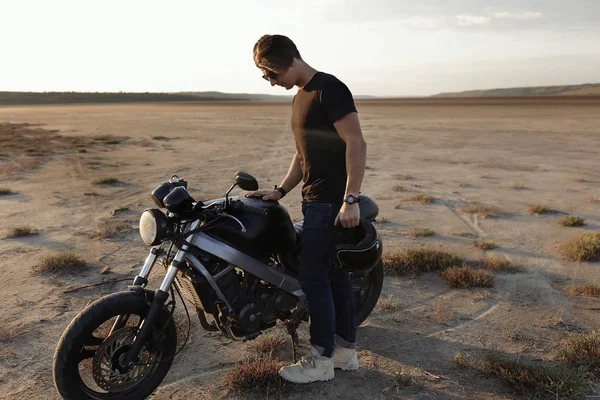 Young man is standig near the motorcycle, in the middle of the desert, helmet with the wings, accessories, sun glasses, sand, tires, risky, free, outdoor, freedom of the soul, looking on bike
