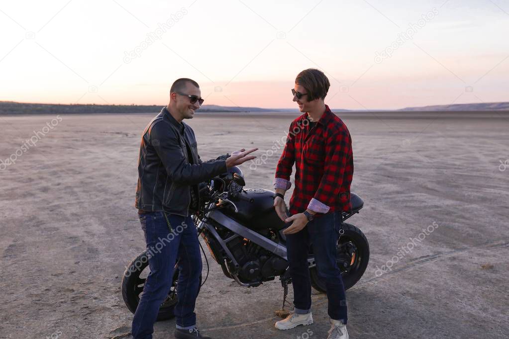 Manly males are standing near the bike in the desert, helmet with the wings, accessories, sun glasses, sand, free, outdoor, freedom of the soul,confident, amazing view, adventure, sunset, having fun  