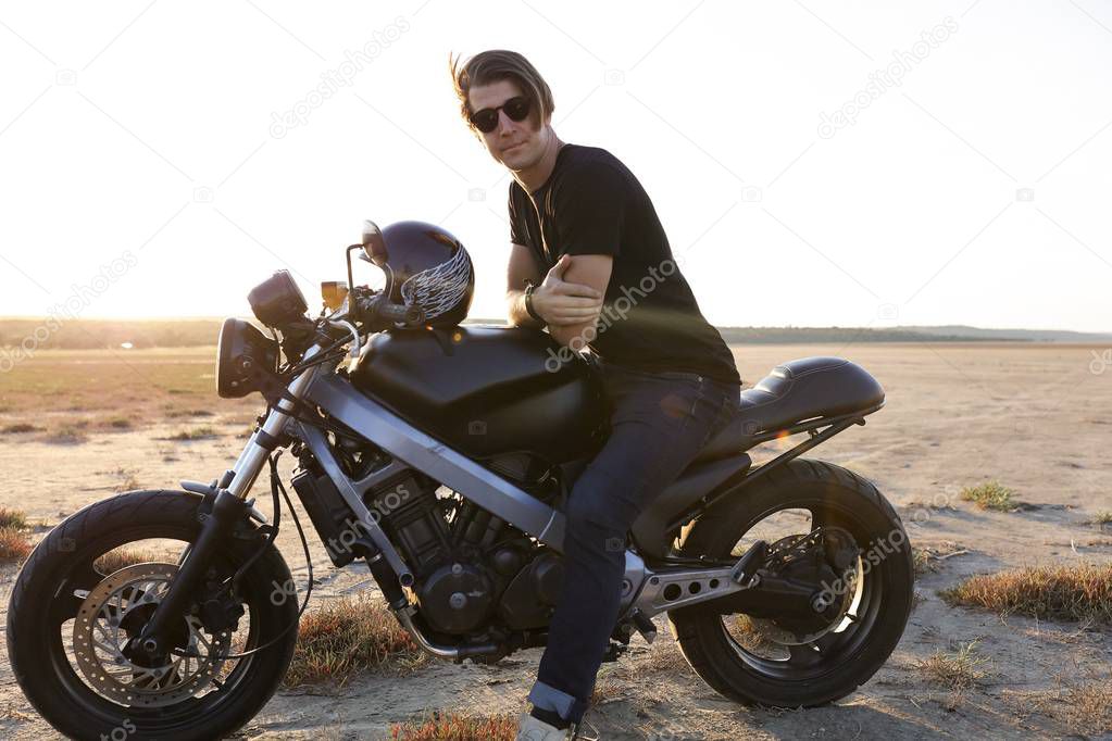Confident risky guy sitting on  the motorbike, in the middle of the desert, helmet with the wings, accessories, sun glasses, sand, tires, free, outdoor, freedom of the soul, sunset, jeans