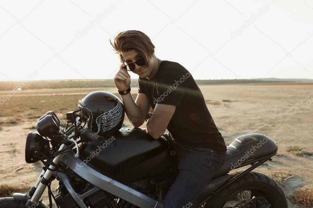 Attractive young guy sitting on  the motorbike, in the middle of the desert, helmet with the wings, accessories, sun glasses, sand, tires, risky, free, outdoor, freedom of the soul, sunset