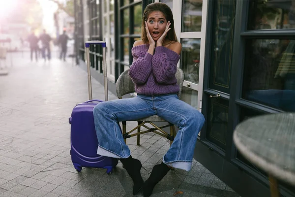 Attractive woman with the suitcase sitting on the chair near the cafe, screaming, wow, in the purple pullover, jeans, makeup, emotions, surprised, autumn, blonde, knitted, boots with the hills