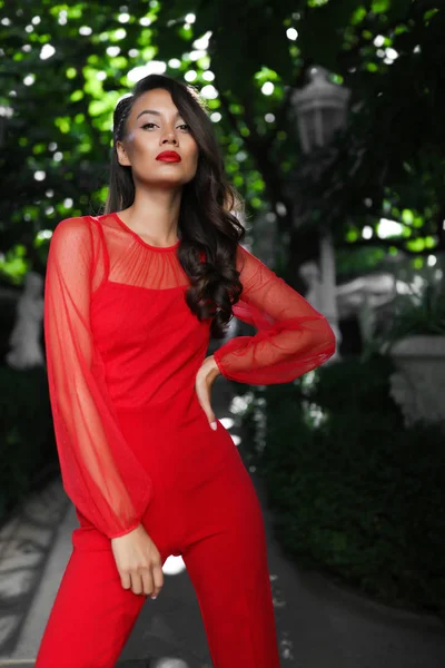 Foxy brunette model in the red suit standing in the garden, hair style, curly,  makeup, heels, shoes, blouse, pants, bright, lady, glamorous, fashion, hand on the waist