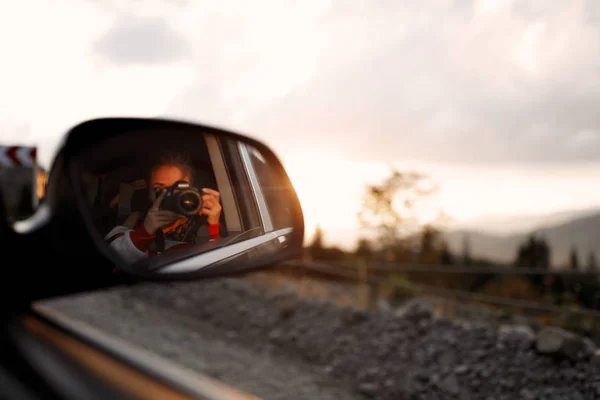 Hipster girl in the car making a photos , mirror, background landscape, amazing view, sunset, road, travelling, calm, joy, camera