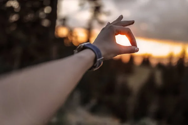 Woman\'s hand on the sunset background, forest, trees, outdoor, watches on the arm, accessories, clouds, rocks, mountains, everything is good