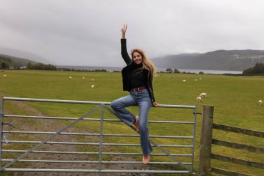 Genç gülümseyen kadın siyah ceket ve kot pantolon, ahşap kapı sheepfold, inanılmaz arka plan görünüm, göl, dağlar, ağaçlar, kırsal, koyunları, yanında oturan yeşil çimen