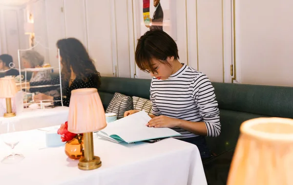 Sweet girl in the striped shirt sitting on the sofa in the restaurant, reading a book, glasses, table lamp, warm, red lips, alone