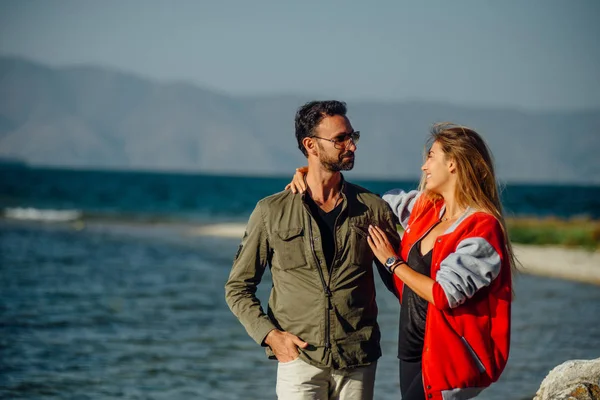 站在海岸上的一对可爱的恋人 认真的家伙在墨镜和绿色衬衫 穿着红色夹克的年轻女士跟他说话 周围的自然 — 图库照片