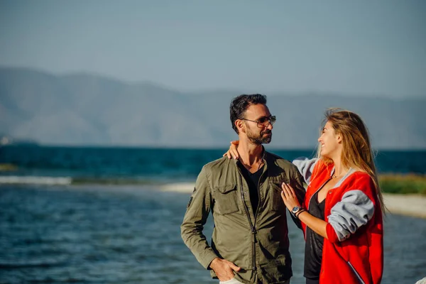 Cute couple standing on the beach near the sea. Hugging and talking to each other. Pretty blonde flirting with the male in the sunglasses. Mountains on the background.