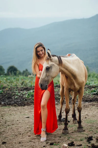 赤いドレスとブロンドの髪でゴージャスな女性 フィールドの馬の近くに立っています 野性の周り 環境との調和 — ストック写真