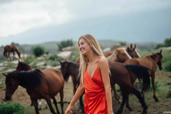 馬と一緒に歩いている女性を笑顔します 美しい金髪 赤いドレスを着ています 栗ホス フィールド放牧します 灰色の空と驚くほど自然風景 — ストック写真