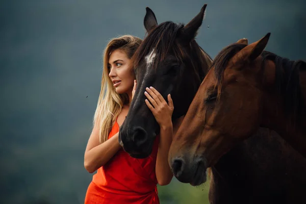 Woman portrait with two brown horses. Young female in the red dress and blonde long hair on the nature background.