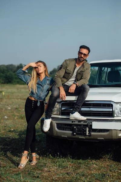 Sweet couple travel by car, road trip. Sexy young blonde and brutal man sitting on the car. Outdoor, beautiful nature landscape.
