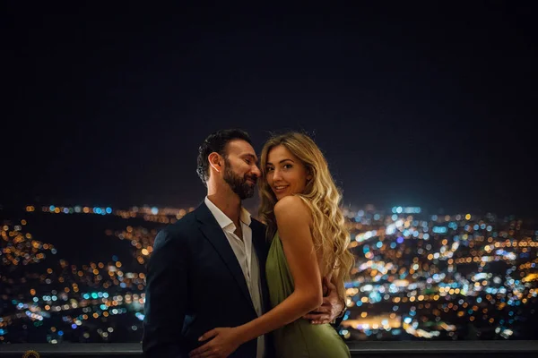 穿着西装的帅哥抱着他豪华的女朋友在露台上 有吸引力的女人在绿色的礼服 化妆和发型 背景上的夜城灯光 — 图库照片