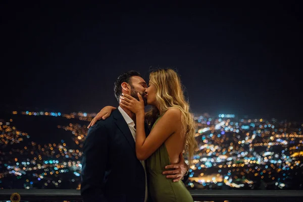 浪漫的气氛 两个人在爱亲吻户外 穿西装的家伙和穿绿色衣服和金发的漂亮女孩 背景上的城市灯光 — 图库照片