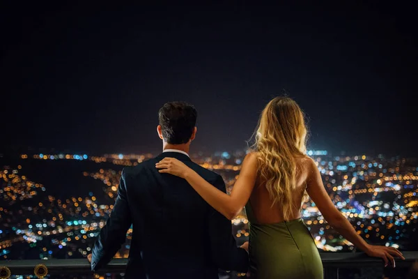 Sweet couple standing together, enjoy the night city view. Man in suit and woman in dress on the celebration.