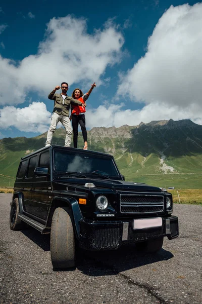 エネルギッシュな男性と女性 車の上に立って 手を振るします 幸せな 笑顔の人々 晴れた日にグルジアの山で — ストック写真