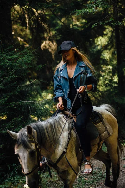 Gorgeous lady riding white horse. Waving blonde hair. Wearing black clothes and jeans jacket. Forest on the background.