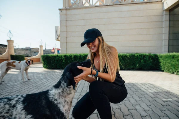Smiling young woman in black pet the dog outdoor. White with red spots dog is on the background. Cheerful blonde loves animals.