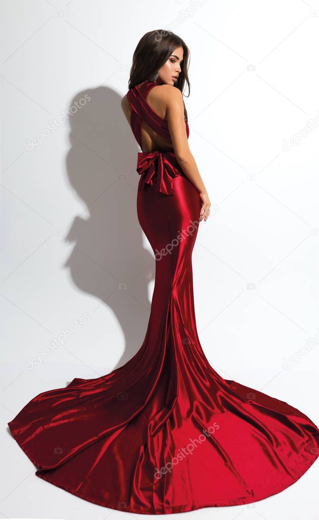 Gorgeous fashion woman in red dress standing. Long hem and bow on the back. Perfect body, makeup and hair style.