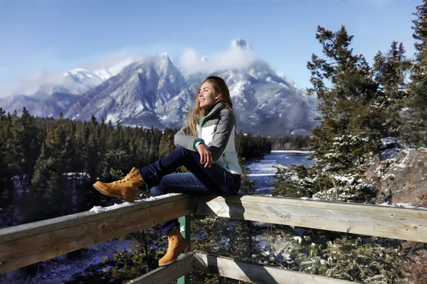 运动女孩坐在木栅栏上 穿着运动服 牛仔裤和靴子 加拿大冬季寒冷的天气 背景上的山脉和雪 — 图库照片