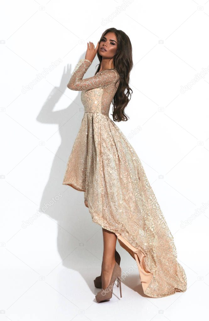 Wonderful lady in creme evening dress standing sideways, hand near face. Curly hair and bright makeup. High heels. Long hem.