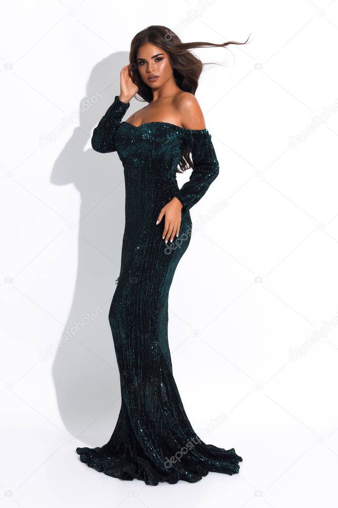 Glamorous woman in dark blue evening dress standing sideways on white background. Waving curly hair, bright makeup and stylish look.