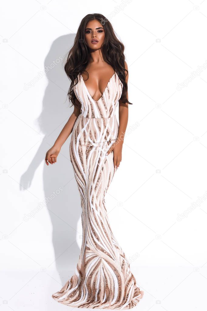 Wonderful sexy female in white long dress with golden design and decollete. Curly volume hairstyle and modern makeup.